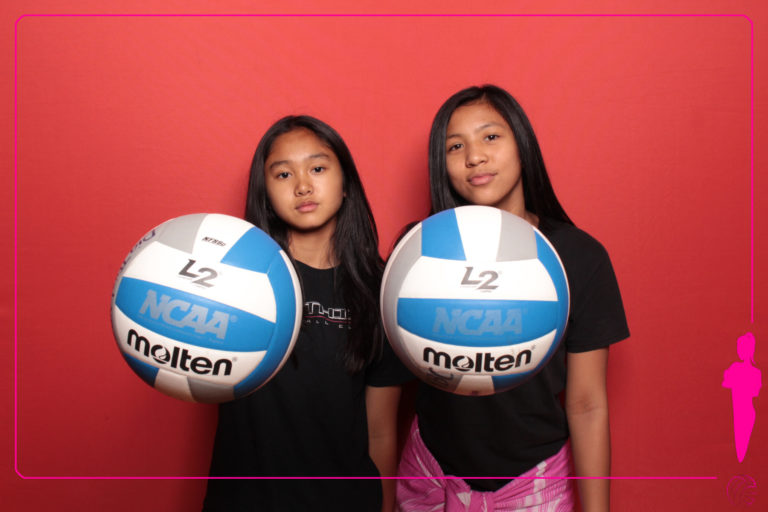 Two women with volleyballs posing in front of red backdrop