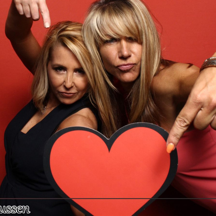 two women posing with red backdrop and heart prop